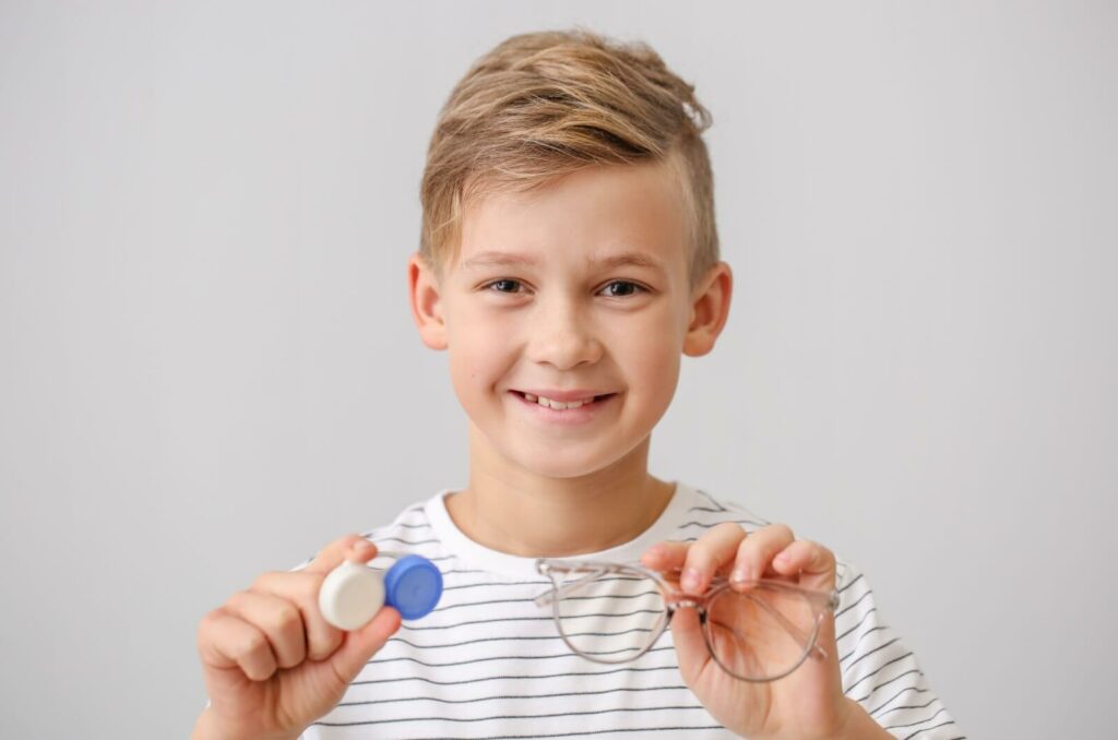 A young boy holding different corrective lenses for myopia, one pair of glasses, and a contact lens case.