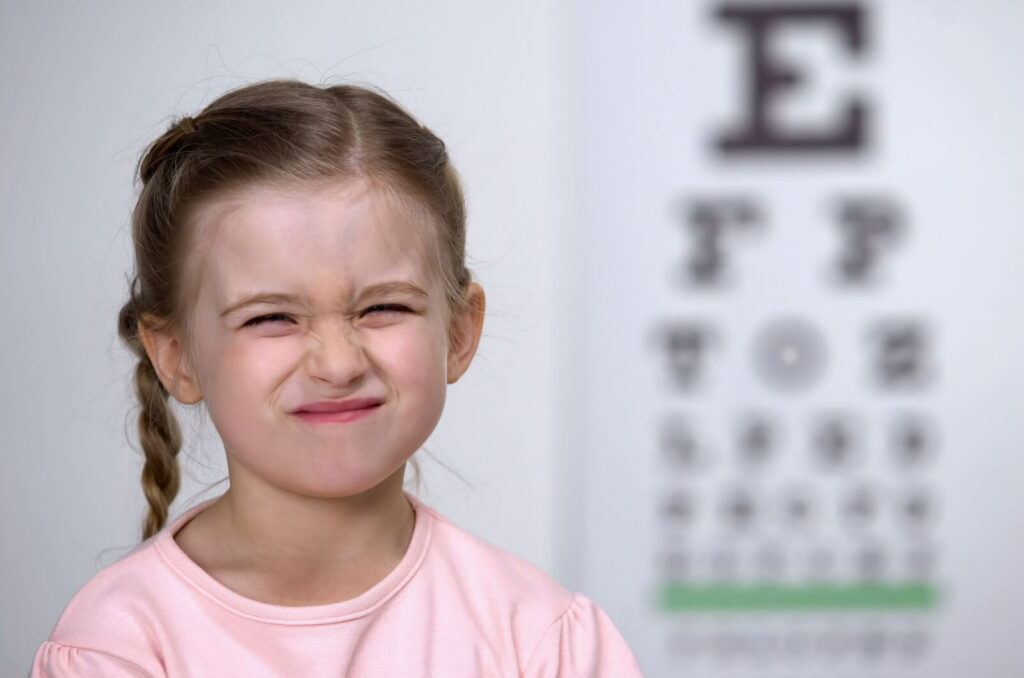 A child squinting in frustration at the camera in front of a blurry letter chart during an eye exam for myopia.