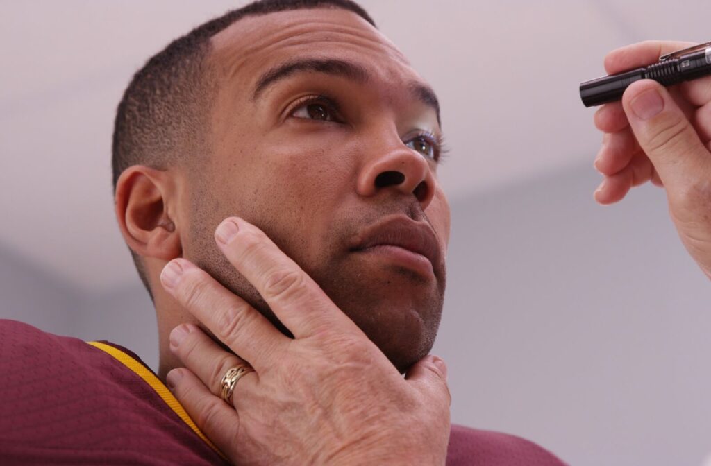 Close-up of a doctor shining a penlight into a man’s eyes during an eye exam, with the doctor’s hand steadying his face.