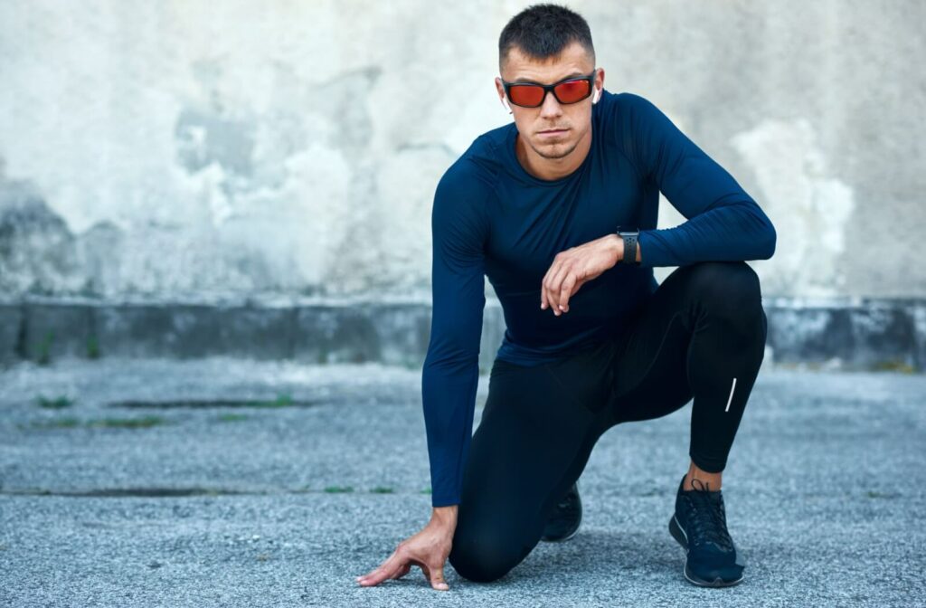 A male athlete in black performance gear kneeling on a track while wearing red-tinted wrap-around prescription sports sunglasses.