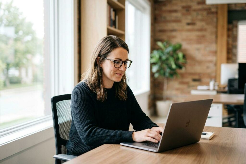 Person in glasses and dark sweater typing on a laptop at a wooden desk by a window, smiling.